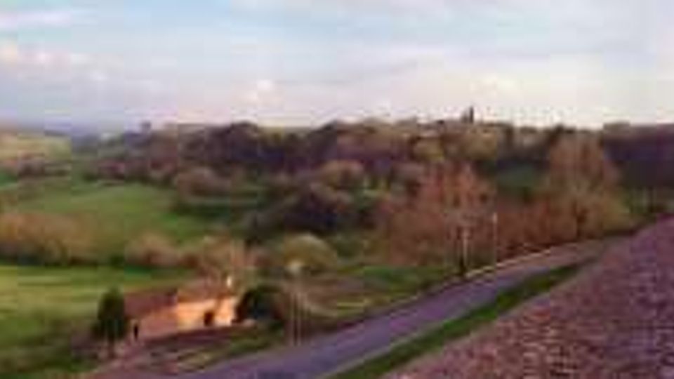 Vista panoramica di una campagna collinare con campi verdi e vegetazione, attraversata da un sentiero in ghiaia lungo un muro in pietra. A destra si vede un prato con vialetti e un edificio storico in pietra con una grande porta ad arco e alberi sullo sfondo.