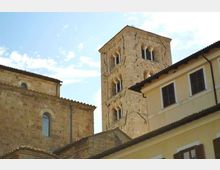 Torre campanaria in pietra chiara con finestre ad arco e colonnine, che svetta tra edifici storici con tetti in coppi e facciate intonacate. Sullo sfondo cielo azzurro con leggere nuvole.