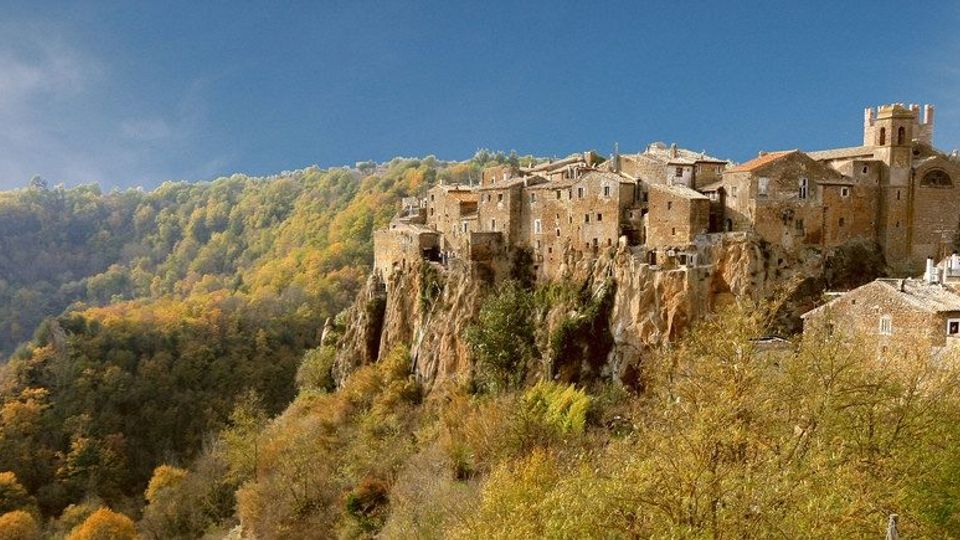 Borgo medievale in pietra arroccato su una rupe, con case addossate l’una all’altra e una chiesa con campanile. Intorno si estende una valle boscosa con colline alberate sotto un cielo azzurro.