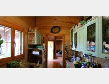 Interno di una cucina rustica in legno con soffitto curvo, finestra a due ante e mobili verde chiaro con ante in vetro. Piano di lavoro con utensili e oggetti, pareti decorate con piastrelle e foto, e porta che conduce a un corridoio con pavimento in piastrelle.