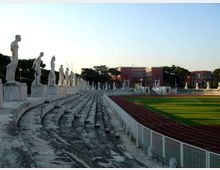 Gradinate in pietra di uno stadio all’aperto con una fila di statue classiche su piedistalli lungo il lato sinistro. A destra si vedono la pista di atletica rossa e il campo verde recintato, con edifici sullo sfondo sotto un cielo sereno.