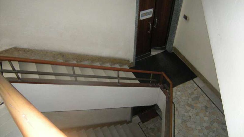 Vano scala interno di un edificio, visto dall’alto, con gradini in cemento, corrimano in legno e ringhiera metallica. Sul pianerottolo si vedono pareti chiare, pavimento in pietra/mosaico e una porta scura con cornice in marmo.