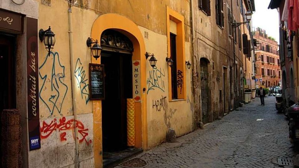 Vicolo urbano stretto con pavimentazione in sampietrini, fiancheggiato da edifici storici intonacati con finestre a persiane e lampioni a parete. In primo piano un ingresso ad arco con cornice arancione su una facciata con graffiti, mentre la strada prosegue tra palazzi fino a un incrocio sullo sfondo.