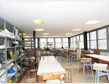 Aula laboratorio artistico con grandi tavoli da lavoro e sgabelli, scaffalature laterali piene di materiali e oggetti. Ampie finestre lungo una parete illuminano l’ambiente, con colonne scure e lampade fluorescenti a soffitto.