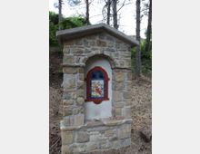 Piccolo tabernacolo votivo in pietra con tetto a spiovente e nicchia ad arco, contenente un’edicola dipinta incorniciata in rosso e blu. È situato su una scarpata in un’area boschiva con pini e sottobosco.