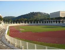 Stadio all’aperto con pista di atletica rossa che circonda un campo in erba, gradinate in pietra e recinzione metallica lungo il bordo. Sullo sfondo si vedono una fila di statue e alberi, una collina verde e un edificio rettangolare a più piani.