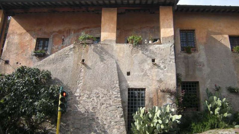 Facciata di un edificio rustico in intonaco e pietra con un grande contrafforte centrale, finestre con inferriate e tetto a falde sporgente. In primo piano vegetazione con cactus e un piccolo semaforo su palo giallo, con muretto di pietra ai bordi.