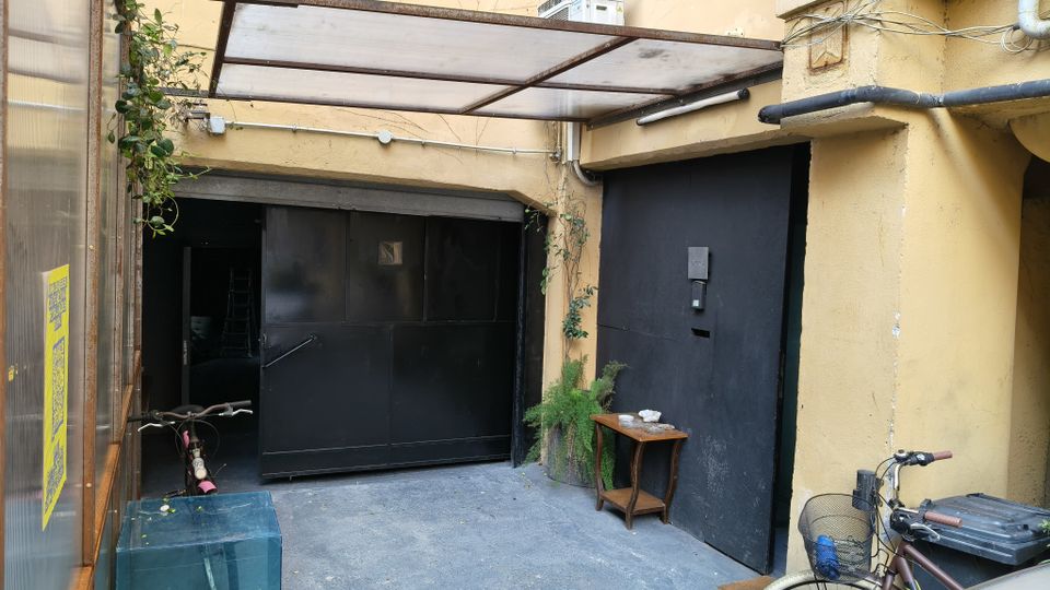 Cortile interno di un edificio con pavimento in cemento e pareti color ocra, coperto da una tettoia in metallo e pannelli traslucidi. Si vedono grandi portoni neri, tubazioni a vista, piante rampicanti e una piccola mensola in legno vicino all’ingresso.