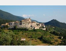 Borgo medievale su una collina con case in pietra addossate e una chiesa con campanile al centro, circondato da mura e terrazzamenti. Sullo sfondo si vedono montagne boscose e una valle verde sotto un cielo sereno.