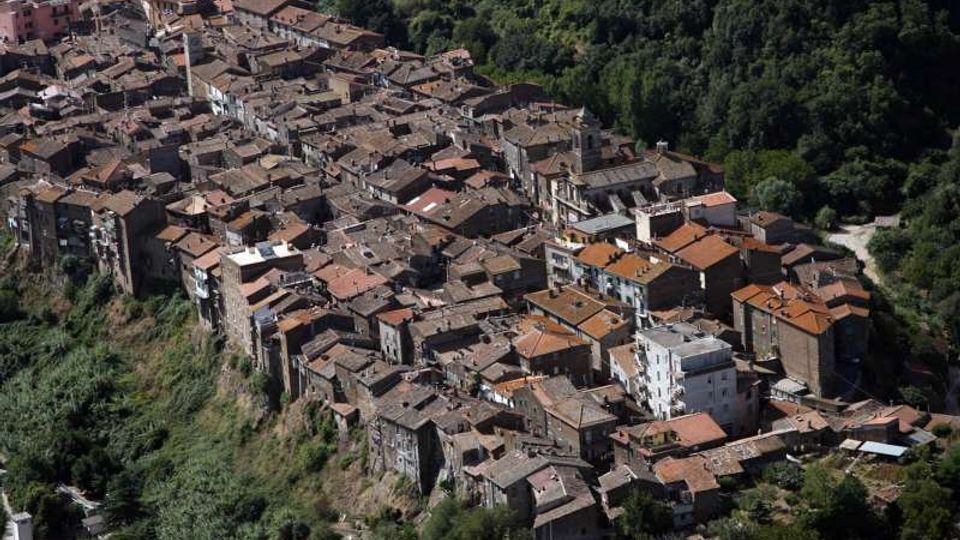 Vista aerea di un borgo collinare con case in pietra addossate tra loro e tetti in coppi, disposto lungo un crinale. Il centro abitato è circondato da fitta vegetazione e pendii boscati.