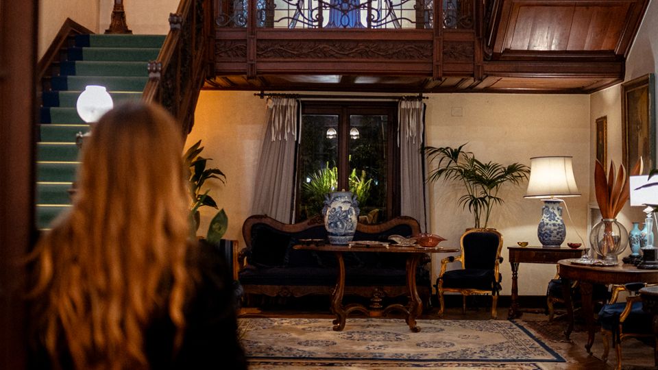 Interno di un salone in stile classico con soppalco in legno e ringhiera in ferro battuto, dominato da una grande vetrata decorativa. A sinistra una scala con moquette verde, e al piano terra divani e tavolini con lampade e vasi su tappeti orientali, davanti a una finestra con tende.