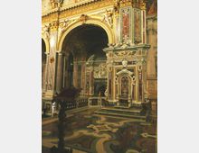 Interno di una chiesa barocca con grandi archi dorati e colonne, pareti riccamente decorate con marmi policromi, stucchi e statue. In primo piano si vede un pavimento in marmo intarsiato e un altare laterale ornamentale con pannelli scolpiti e decorazioni elaborate.