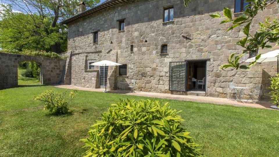 Cortile erboso con arbusti davanti a un edificio rurale in pietra a due piani, con piccole finestre e porte vetrate che si aprono su un patio. Sulla terrazza ci sono tavoli e sedie con ombrelloni bianchi, e a sinistra un arco in pietra conduce a un’area verde.