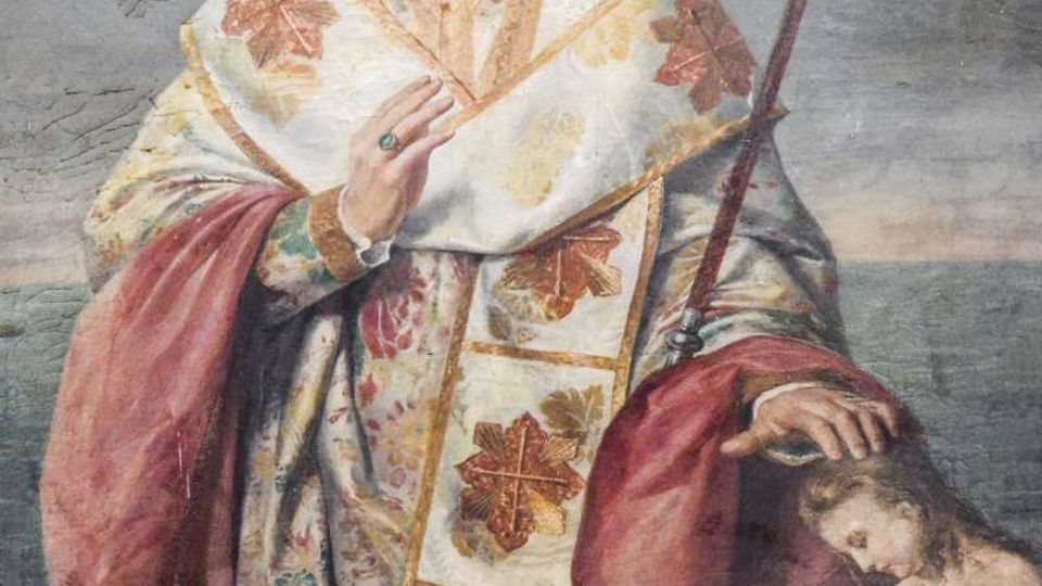 Affresco religioso su parete in pietra raffigurante un santo con aureola e vesti liturgiche riccamente decorate, con pastorale e mano alzata in gesto di benedizione. Ai suoi piedi è visibile una piccola scena con figure infantili, dipinta nello stesso riquadro murale.