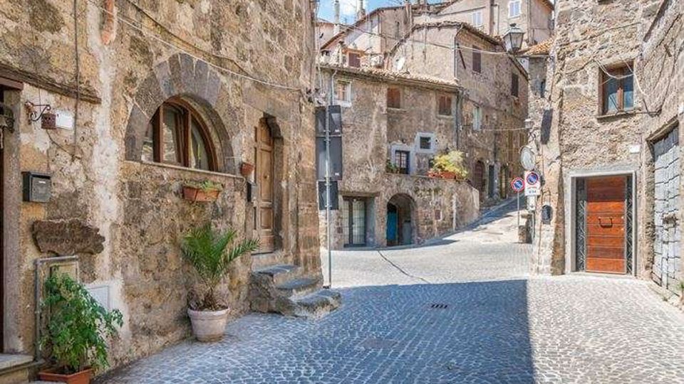 Strada acciottolata in un borgo storico, fiancheggiata da edifici in pietra con archi, finestre piccole e porte in legno. La via curva e sale leggermente tra facciate rustiche con vasi di piante e segnaletica stradale.