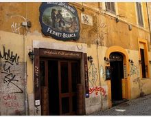Facciata di un edificio storico con intonaco giallo e numerosi graffiti, con insegna vintage “Antico Caffè del Moro – Fernet-Branca” sopra l’ingresso. Due porte ad arco con lanterne a parete si affacciano su una strada lastricata in pietra.