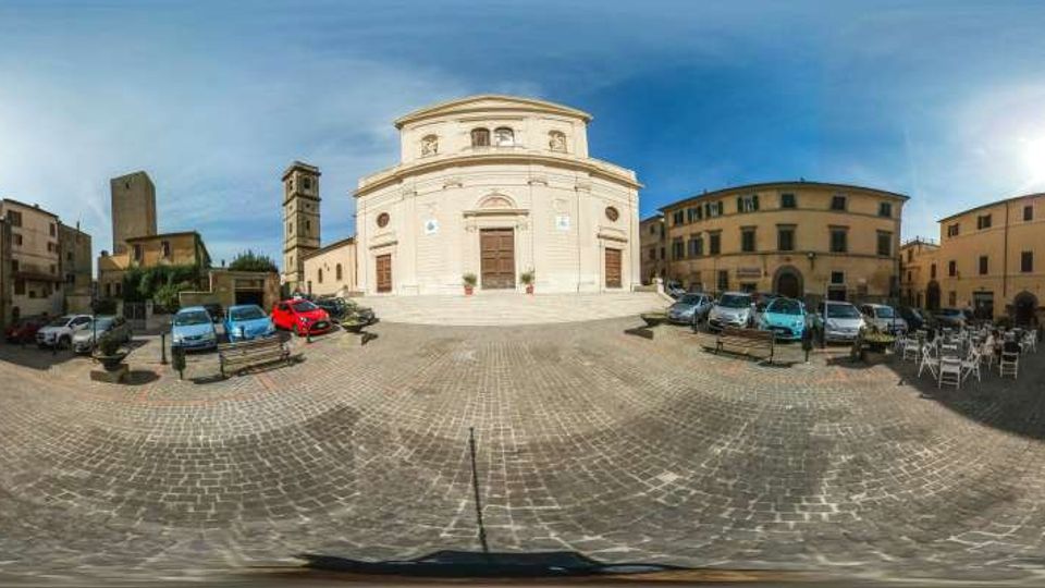 Ampia piazza lastricata in pietra con parcheggi e panchine, dominata al centro da una chiesa in stile neoclassico con facciata curva e portale principale. Intorno si vedono edifici storici a più piani e un campanile, sotto un cielo sereno.