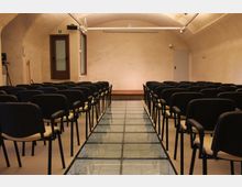 Sala conferenze con soffitto a volta e file di sedie nere disposte ai lati di un corridoio centrale con pavimentazione in vetro. In fondo si vedono una parete chiara, una porta e illuminazione a binario sul soffitto.