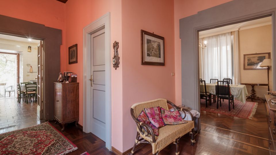 Interno di un appartamento con pareti rosa e pavimento in parquet, con una poltrona in vimini e quadri alle pareti. Due ampie aperture conducono a una zona pranzo con tavolo e sedie su tappeto e a un’altra stanza con tavolino e porta finestra.
