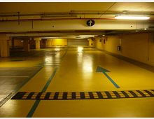 Parcheggio sotterraneo con pavimento giallo e linee blu di corsia, con una grande freccia direzionale dipinta a terra e un dosso rallentatore in primo piano. Soffitto basso con tubazioni e illuminazione al neon, colonne e pareti gialle lungo le corsie.