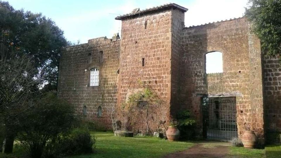 Edificio storico in pietra con mura spesse e una torre centrale, affiancato da un arco d’ingresso con cancello metallico. Davanti si vede un cortile erboso con un sentiero, cespugli, alberi e grandi vasi in terracotta.