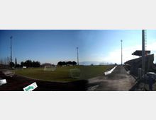 Campo da calcio all’aperto con pista di atletica in asfalto scuro, porte e piccoli tabelloni pubblicitari lungo il bordo. A destra si vedono tribune coperte, alti pali per l’illuminazione e una recinzione, con alberi e cielo sereno sullo sfondo.