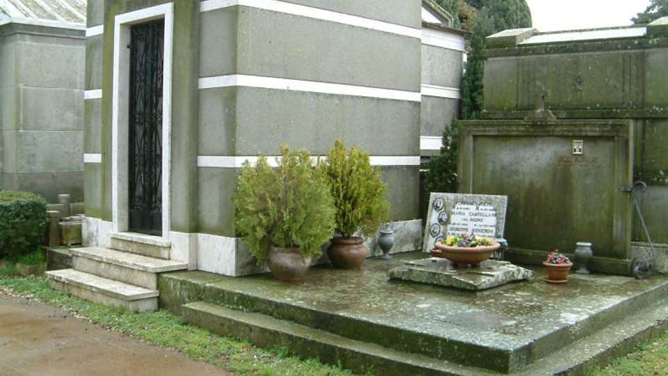 Cappella funeraria in un cimitero, con edificio in muratura grigia a fasce bianche e porta metallica raggiunta da pochi gradini. Davanti c’è una piattaforma in cemento con lapide, vasi di fiori e piccoli arbusti, circondata da altri loculi e cipressi sullo sfondo.
