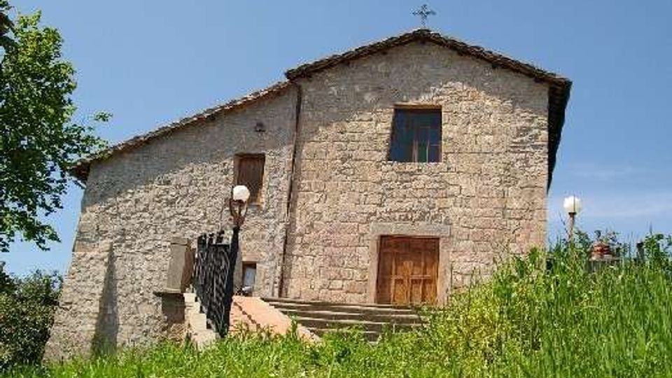 Edificio storico in pietra, simile a una piccola chiesa, con tetto a falde, porta in legno e una croce sulla sommità della facciata. L’ingresso è raggiungibile tramite una scalinata con ringhiera, circondato da erba alta e alberi sotto un cielo sereno.