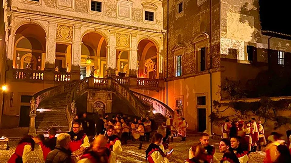 Palazzo storico illuminato di notte con loggiato ad archi e ampia scalinata centrale che conduce al piano superiore. In primo piano si vede una piazza lastricata in pietra, circondata da facciate antiche.