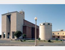 Chiesa moderna con facciata in pietra chiara e volumi geometrici, affiancata da una struttura cilindrica e da una torre campanaria con croce. Davanti si vede una piazza pavimentata con aiuole, piccoli alberi e un lampione, sotto un cielo sereno.