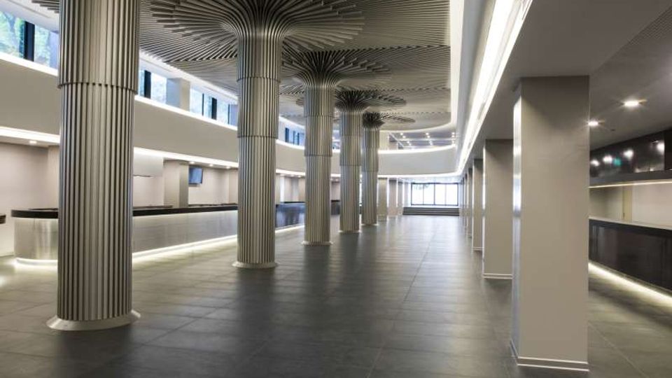 Ampio atrio interno moderno con pavimento in grandi piastrelle scure e banconi lineari lungo i lati. Colonne cilindriche scanalate sostengono un soffitto con elementi radiali decorativi, illuminate da strisce LED e finestre alte lungo il perimetro.