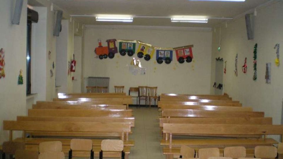 Sala polifunzionale con file di lunghe panche e sedie in legno disposte verso una parete frontale. Soffitto con luci al neon, finestre laterali e decorazioni a tema trenino appese alla parete di fondo.