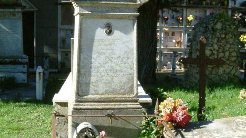 Monumento funerario in marmo in un cimitero, con croce in cima e lapide incisa su un basamento rialzato. Intorno si vedono prato, fiori e altre tombe con loculi sullo sfondo, parzialmente ombreggiate da alberi alti.