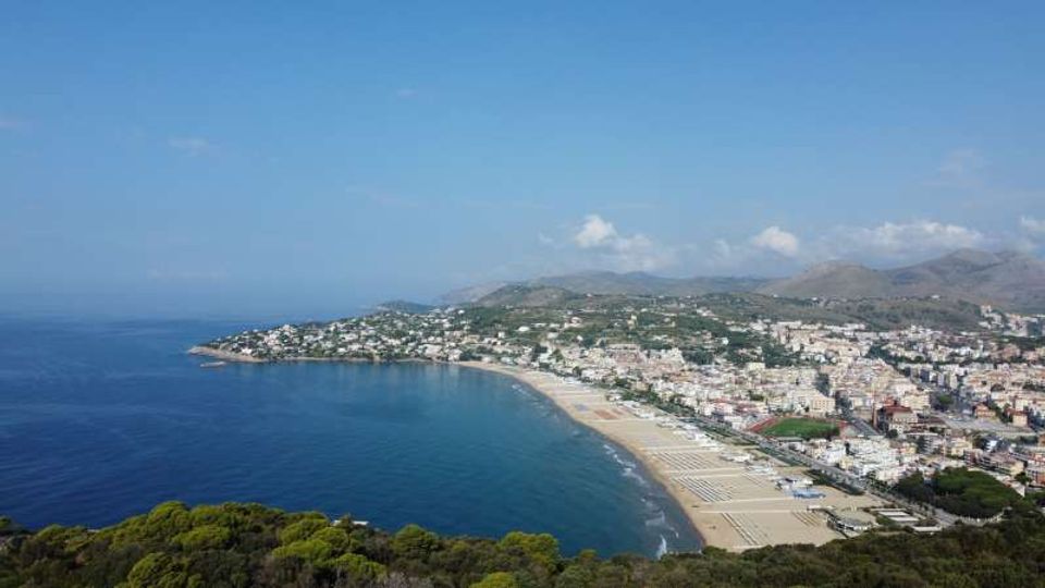 Veduta panoramica dall’alto di una grande baia costiera con lunga spiaggia sabbiosa e mare calmo, affiancata da un centro abitato denso lungo il litorale. Sullo sfondo si vedono colline e montagne, con vegetazione in primo piano e cielo sereno.