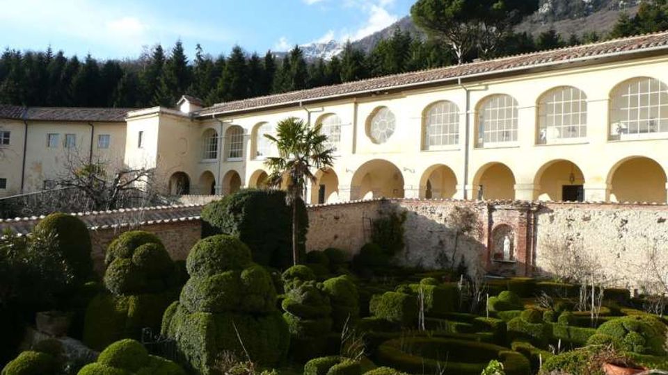 Cortile di un complesso storico con porticato ad archi e grandi finestre ad arco, affacciato su un giardino formale con siepi potate e vasi in terracotta. Sullo sfondo si vedono alberi, montagne rocciose e cielo parzialmente nuvoloso.