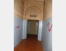 Corridoio interno stretto con pavimento in piastrelle rosse e pareti rivestite da piastrelle azzurre nella parte inferiore e intonaco beige sopra. In fondo c’è un’apertura con porta e un soffitto alto con arco e lampada sospesa, con alcune scritte sui muri.
