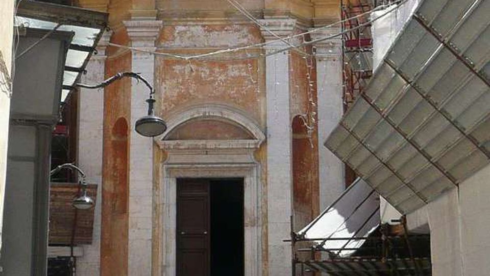 Facciata di una piccola chiesa in stile barocco alla fine di una stretta strada, con scalinata in pietra che conduce al portale e una finestra sopra l’ingresso. Impalcature e teli da cantiere circondano la parte alta dell’edificio e il lato destro della via.