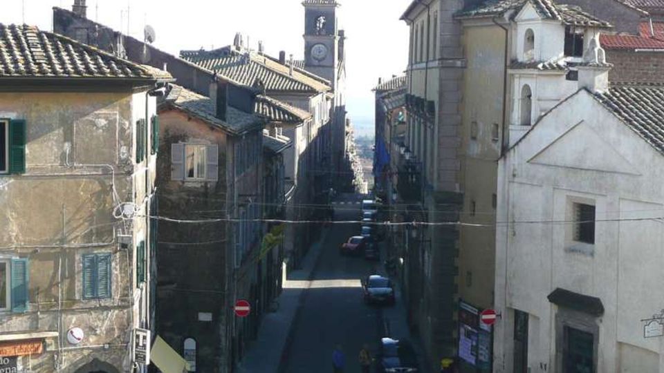 Strada stretta in un centro storico, fiancheggiata da edifici in muratura con tetti in tegole e persiane, con auto parcheggiate lungo i lati. In fondo si vede una torre con orologio che emerge sopra i tetti, mentre la via scende verso l’orizzonte.
