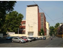Edificio industriale alto con grande torre bianca e struttura laterale in mattoni rossi con finestre verticali, affacciato su una strada urbana. In primo piano ci sono un parcheggio con molte auto e binari del tram con cavi aerei, circondati da alberi.