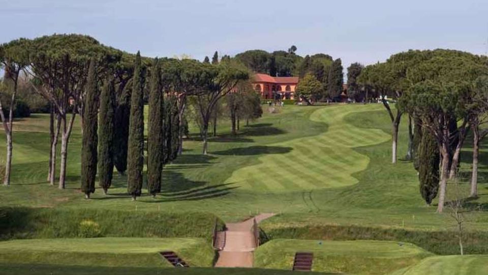 Campo da golf con ampi fairway ondulati a strisce, delimitato da cipressi e pini mediterranei. In primo piano si vede un piccolo ponte sul percorso e sullo sfondo un edificio con tetto rosso tra gli alberi.