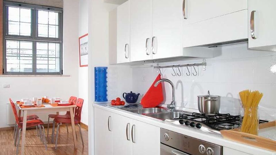 Cucina moderna e luminosa con mobili bianchi, piano cottura a gas con forno in acciaio, lavello e ganci a parete sopra il piano di lavoro. Sullo sfondo c’è un angolo pranzo con tavolo apparecchiato e sedie rosse vicino a una grande finestra.