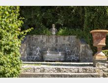Fontana in pietra in un giardino, con vasca rettangolare e mascherone centrale su un muro rivestito in pietra grezza. Sullo sfondo si vede una statua sopra il muro, circondata da vegetazione fitta, e a destra un grande vaso ornamentale in terracotta su piedistallo.
