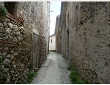 Vicolo stretto con pavimentazione in pietra tra alti edifici in muratura grezza e pietra, con porte e piccole finestre sulle facciate. Il passaggio curva leggermente e conduce verso una casa chiara in fondo, con vegetazione che cresce ai bordi.