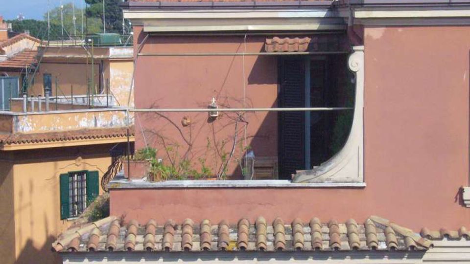 Facciata di un edificio residenziale con pareti rosa e tetto in tegole di terracotta, con una terrazza al primo piano delimitata da parapetto e ringhiera metallica. Sullo sfondo si vedono altri tetti e antenne in un contesto urbano.