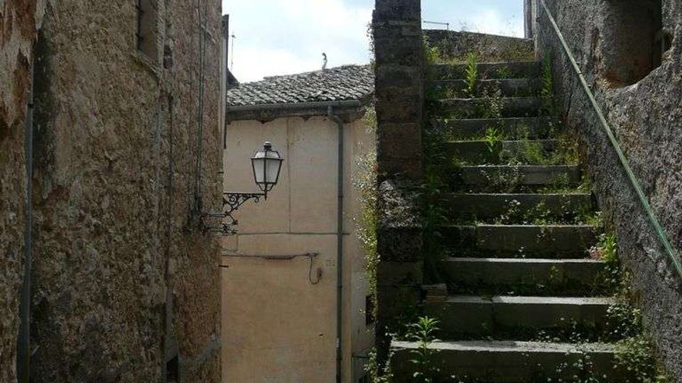 Vicolo stretto tra edifici in pietra con pavimentazione irregolare, illuminato da un lampione a parete. A destra una scalinata in pietra sale tra muretti con vegetazione spontanea, con ringhiera e piccola tettoia in alto.