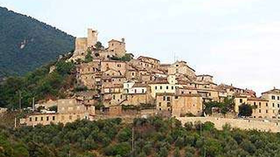 Borgo medievale in pietra arroccato su una collina, con case addossate tra loro e una torre o castello sulla sommità. Sullo sfondo si vedono un pendio montuoso e, in primo piano, una fascia di vegetazione e alberi.