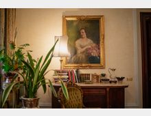 Interno di una stanza in stile classico con grande ritratto incorniciato in oro sopra una console in legno con piano in marmo. Sulla console ci sono una lampada con paralume, libri e oggetti decorativi, con piante in vaso e una sedia imbottita in primo piano.