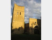 Edificio storico in pietra con alta torre rettangolare e piccole finestre ad arco, affiancato da una struttura più bassa con grande portale ad arco e rosone quadrilobato. Davanti c’è un prato, sullo sfondo cielo nuvoloso.