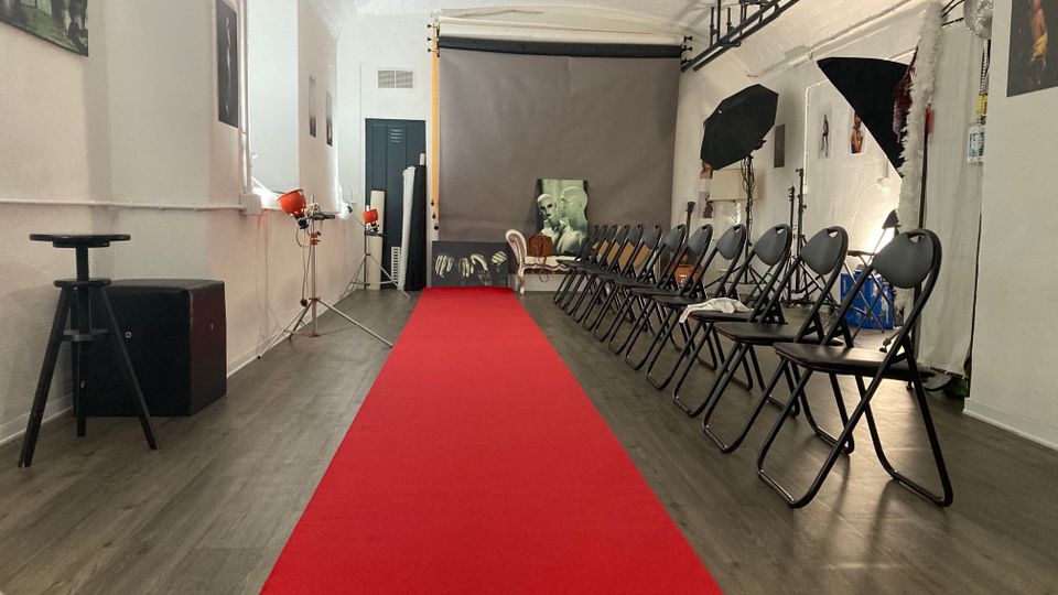Interno di uno studio fotografico con soffitto a volta bianco, tappeto rosso centrale tipo passerella e file di sedie pieghevoli nere lungo il lato destro. Sul fondo si vede un fondale grigio con luci da set, stativi e ombrelli fotografici; alle pareti sono appese alcune immagini.