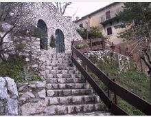 Scalinata in pietra che sale tra muri di roccia verso un edificio in pietra con due finestre ad arco protette da grate. A destra corre una passerella con parapetto in legno, circondata da vegetazione e case con balconi sullo sfondo.